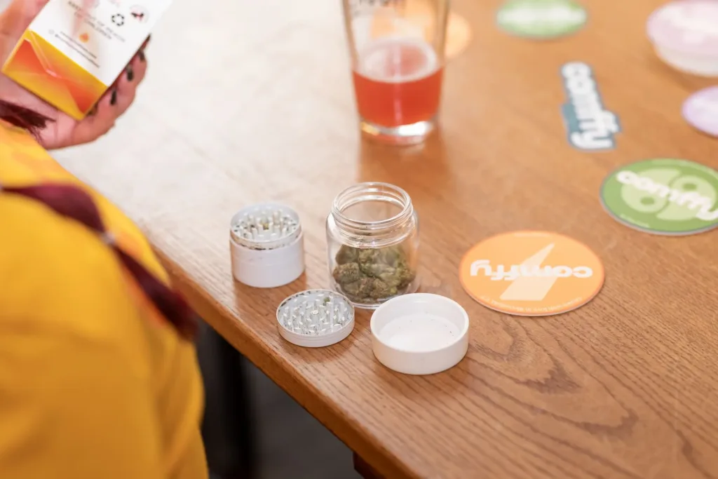 A jar with cannabis buds, a metal grinder, and a drink on a wooden table alongside colorful round coasters.