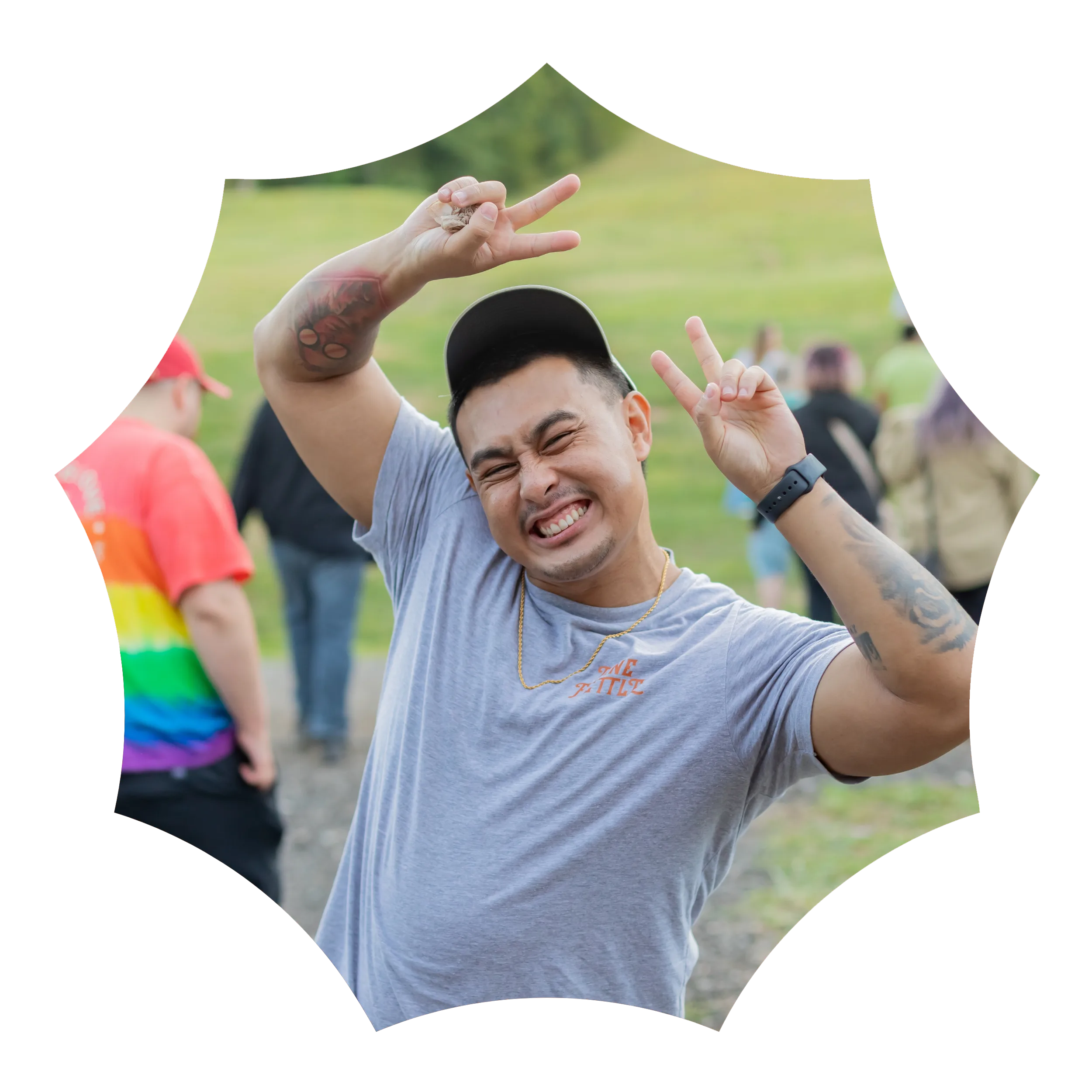 A smiling person in a gray shirt and cap makes peace signs with both hands outdoors, with people and grass in the background.