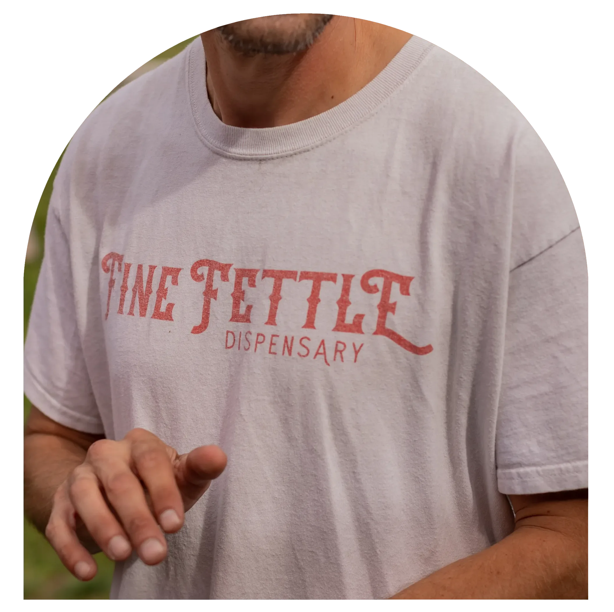 A person wearing a white Fine Fettle cannabis dispensary T-shirt is shown from the shoulders to mid-chest, with one hand partially raised.