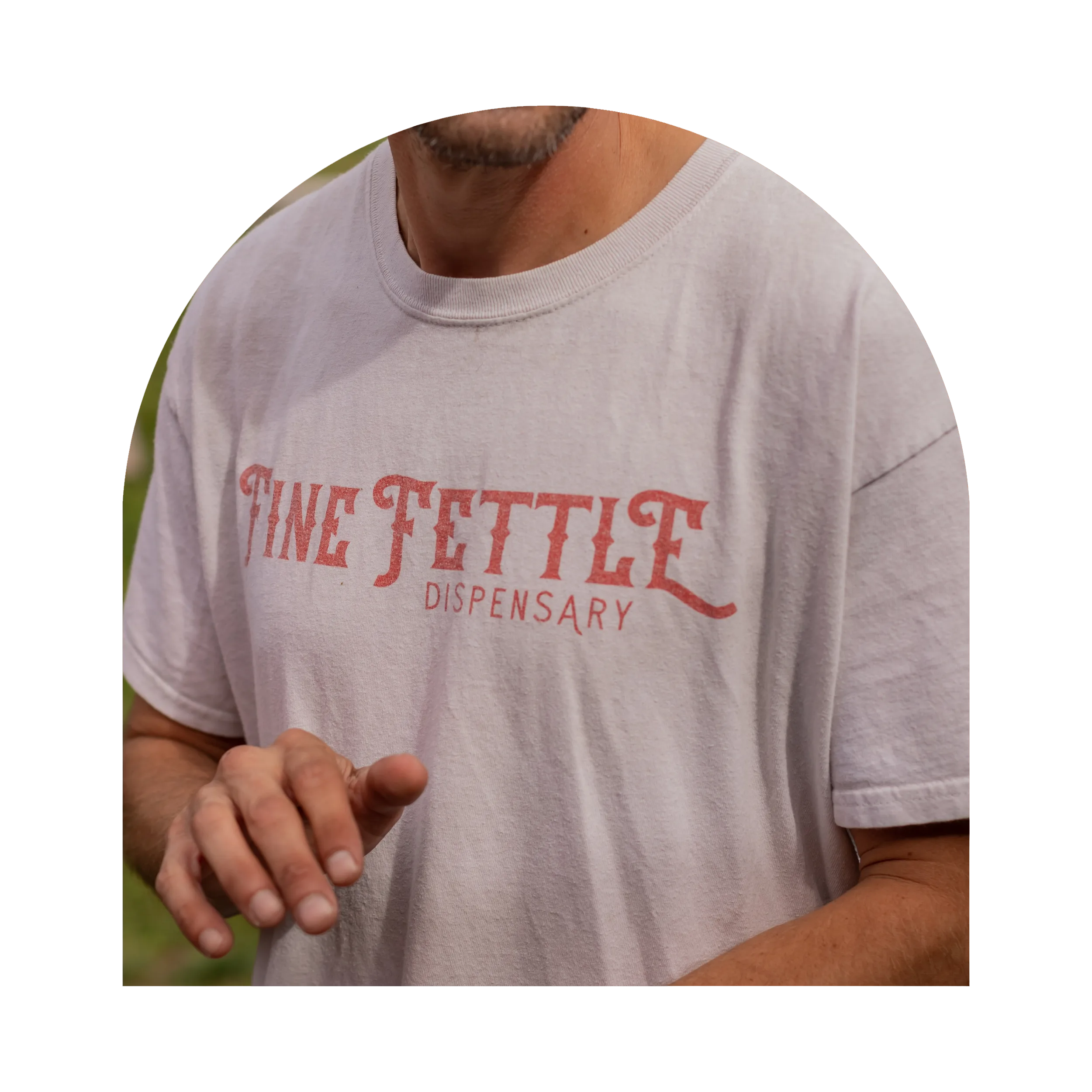 A person wearing a white Fine Fettle cannabis dispensary T-shirt is shown from the shoulders to mid-chest, with one hand partially raised.