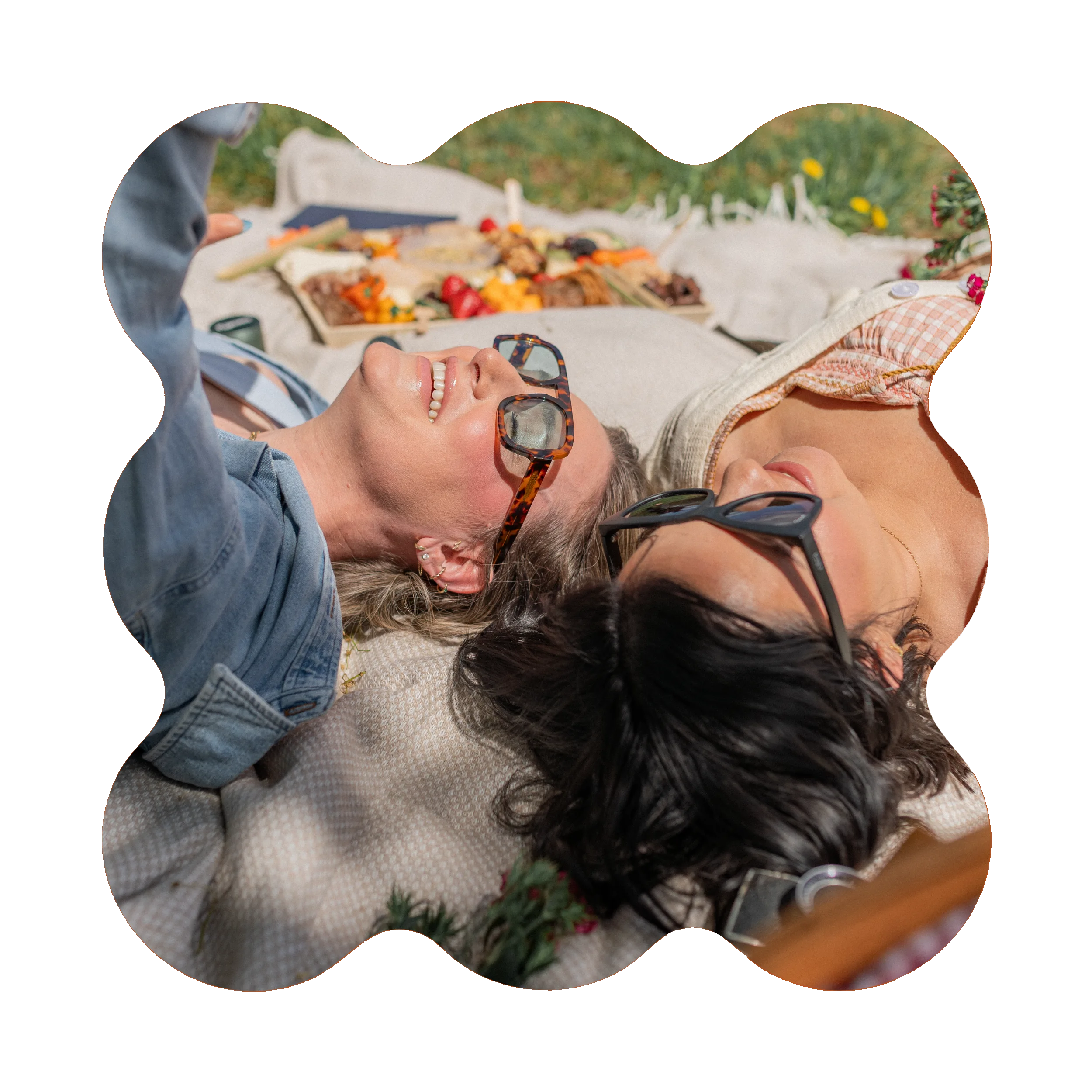 Two women wearing sunglasses lay on a blanket outdoors, enjoying a picnic from a local cannabis dispensary, with food and flowers around them on a sunny day.
