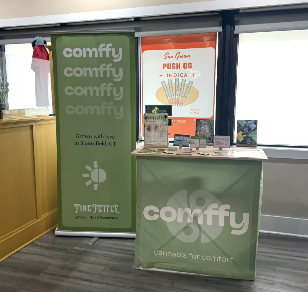 Comffy cannabis display booth with green banners, pamphlets, and a poster. The booth says cannabis for comfort and promotes products grown in Bloomfield, CT.