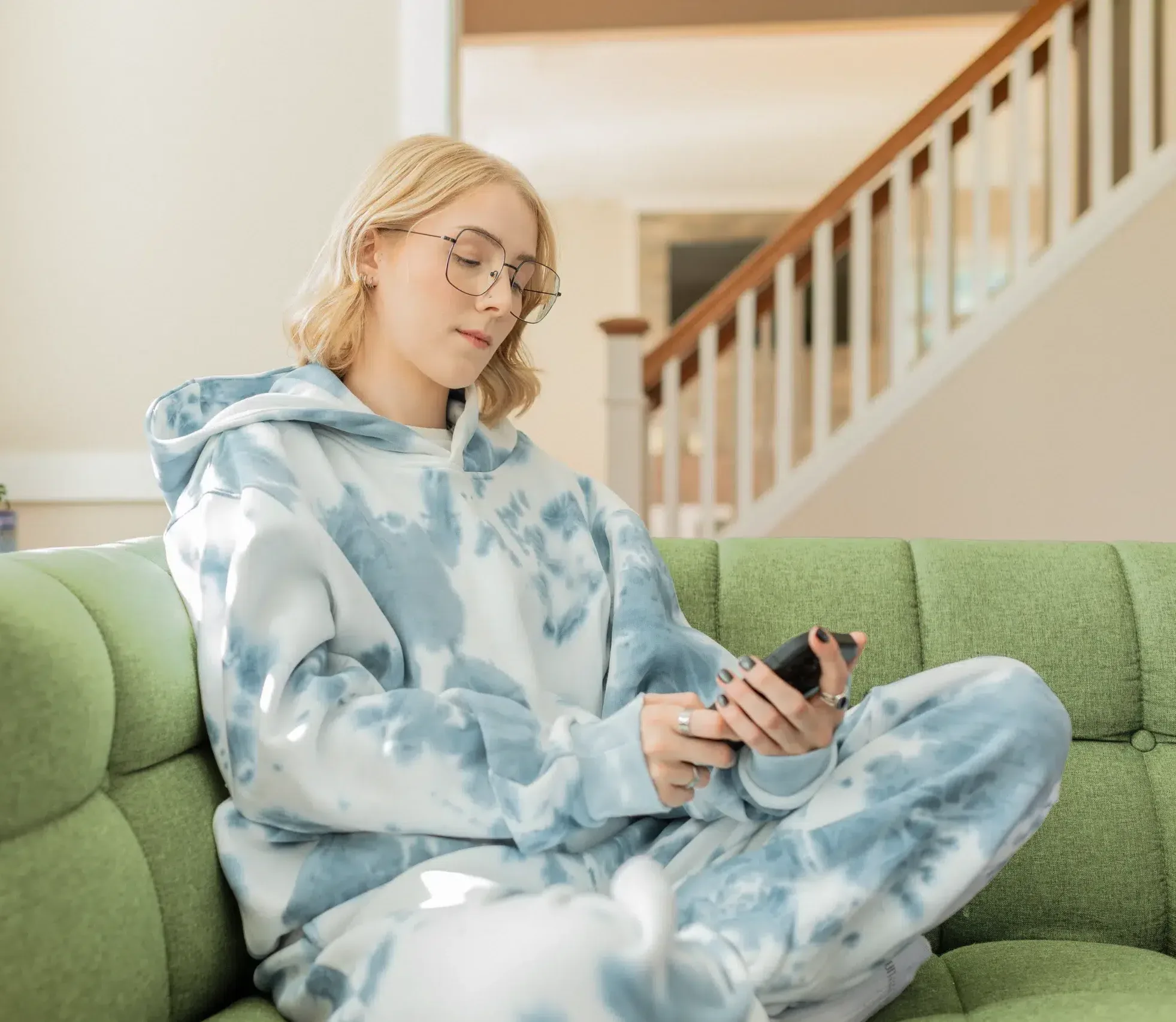 Woman with blonde hair and glasses, wearing a blue tie-dye hoodie and sweatpants, sits on a green couch using her phone. A staircase and potted plant are in the background.