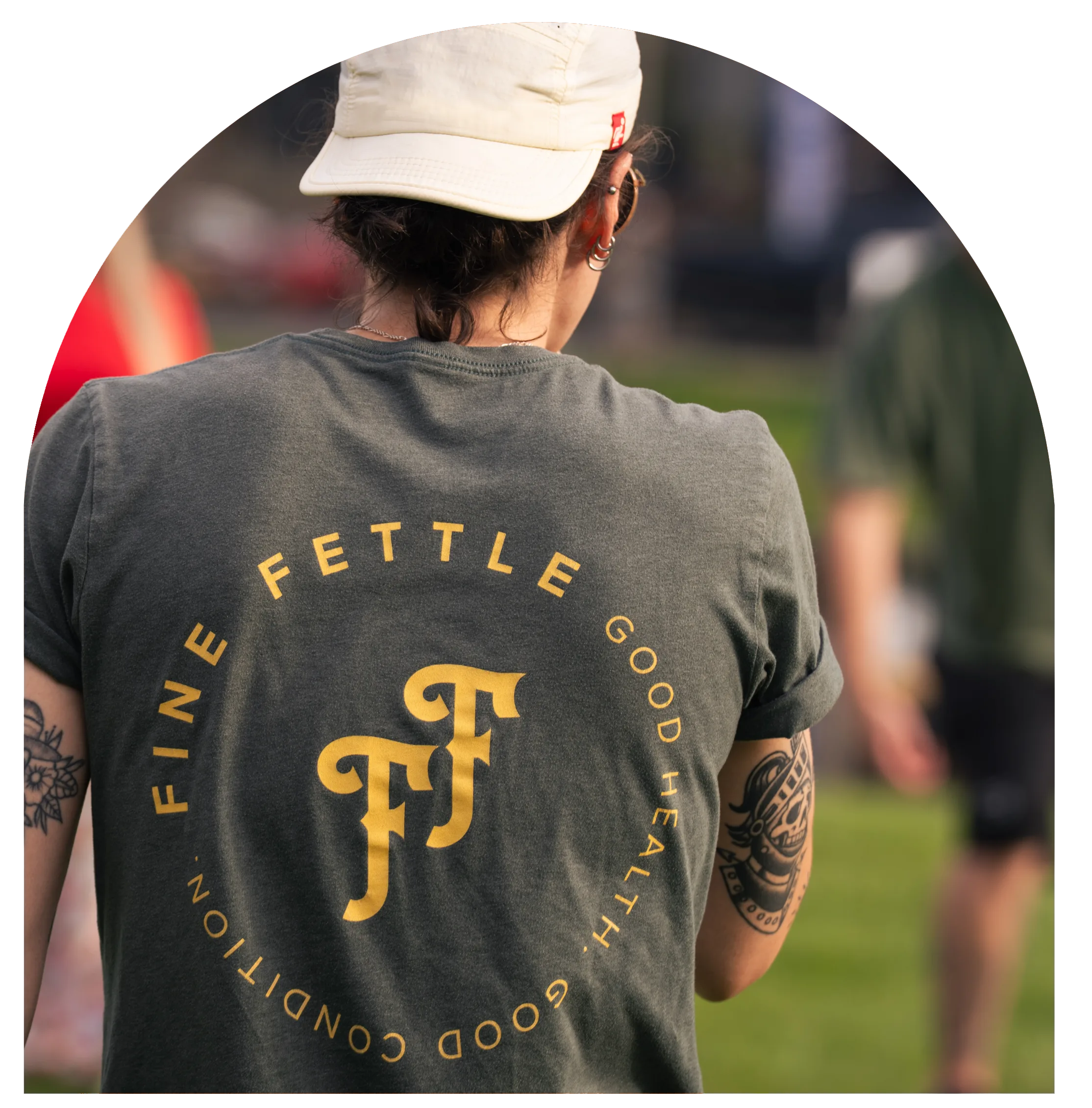 Person in a cap with tattoos wears a shirt reading Fine Fettle Good Health, Good Condition, FF letters—possibly at a CT cannabis dispensary event—outdoors with others in the background.
