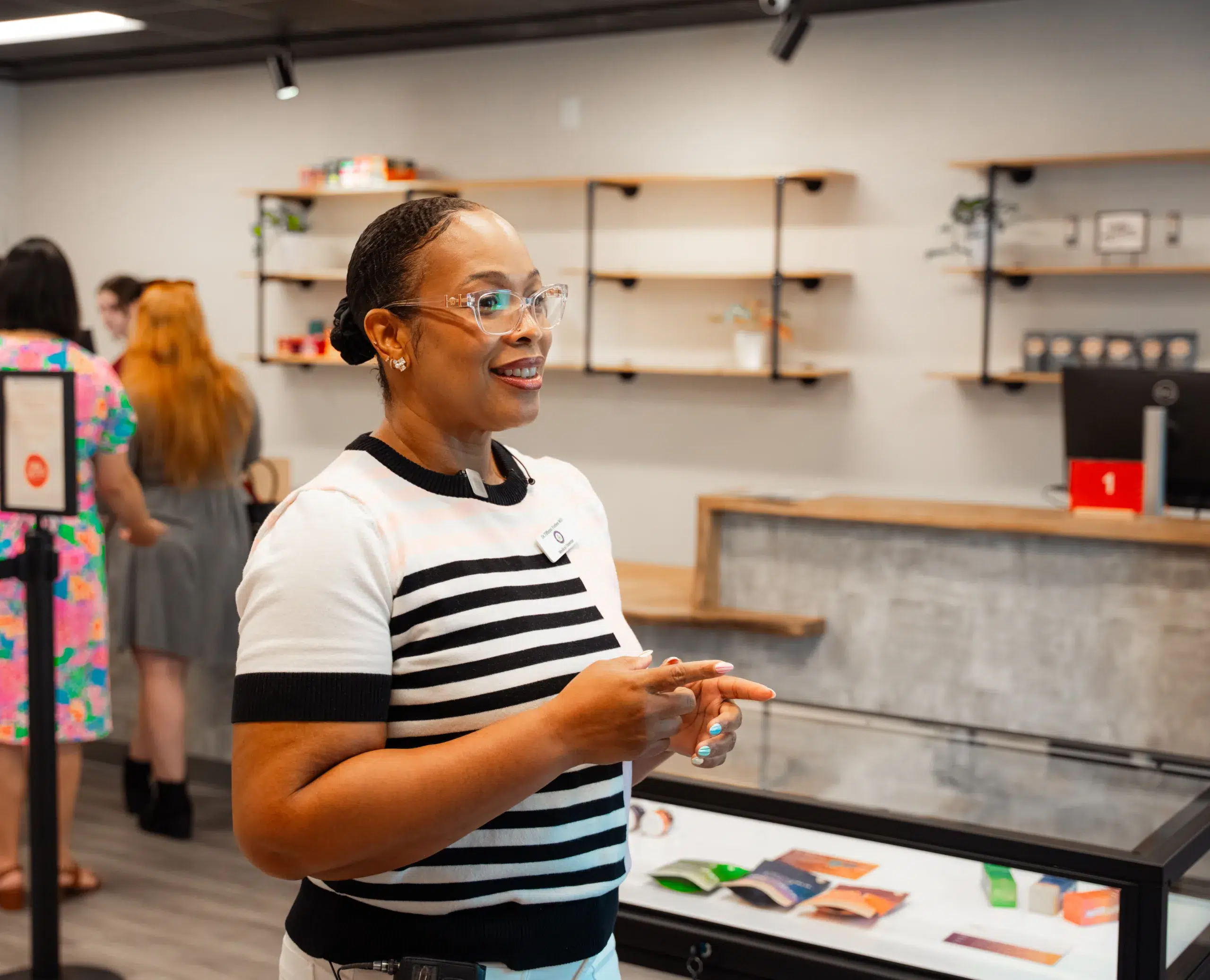 A smiling woman with glasses stands in a modern Georgia dispensary, gesturing with her hands. Shelves and people are visible in the background.