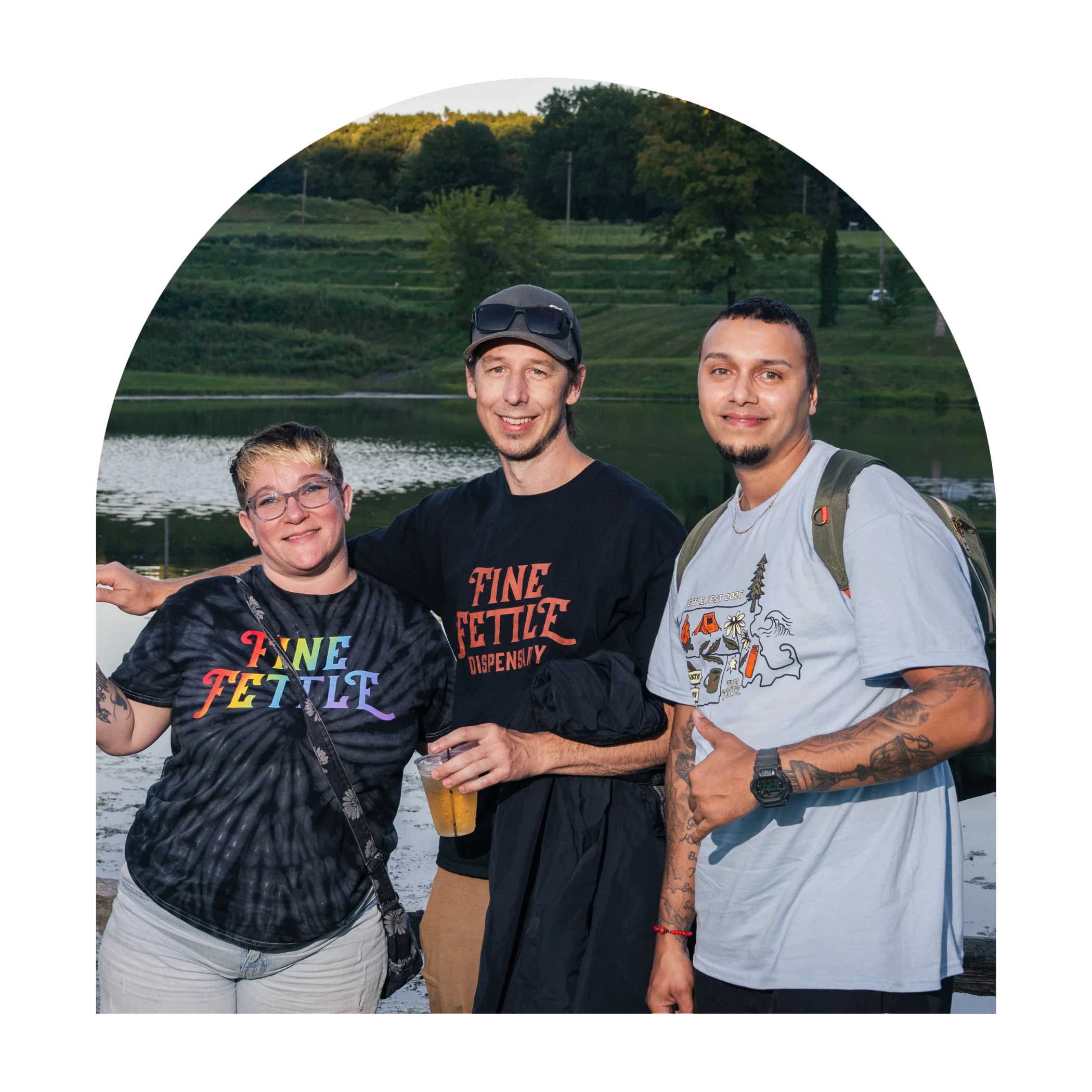 Three people standing in front of a lake; two wear Fine Fettle local dispensary shirts and one gives a thumbs up.