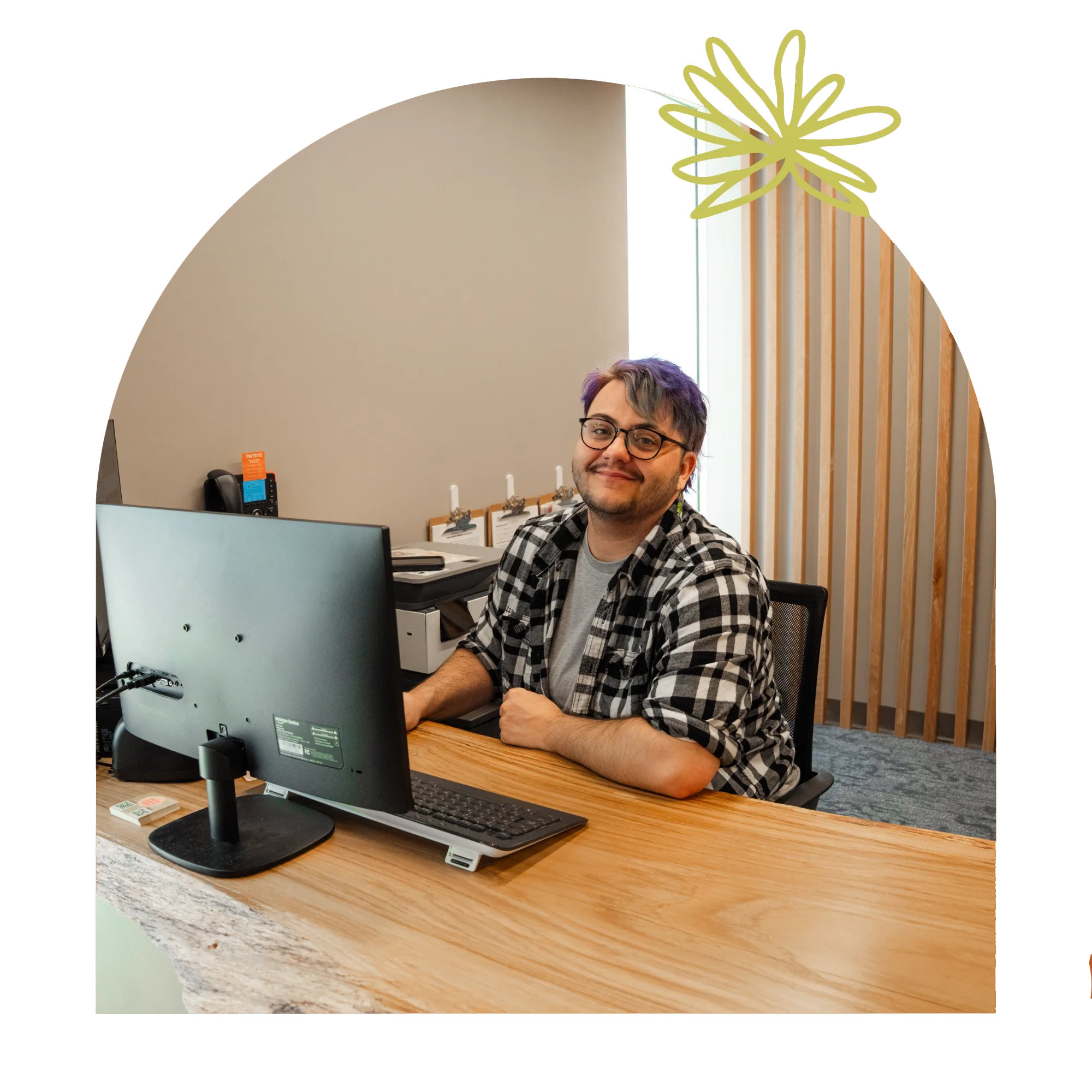 A person with purple-tipped hair and glasses sits at a desk with a computer, working for a local dispensary.