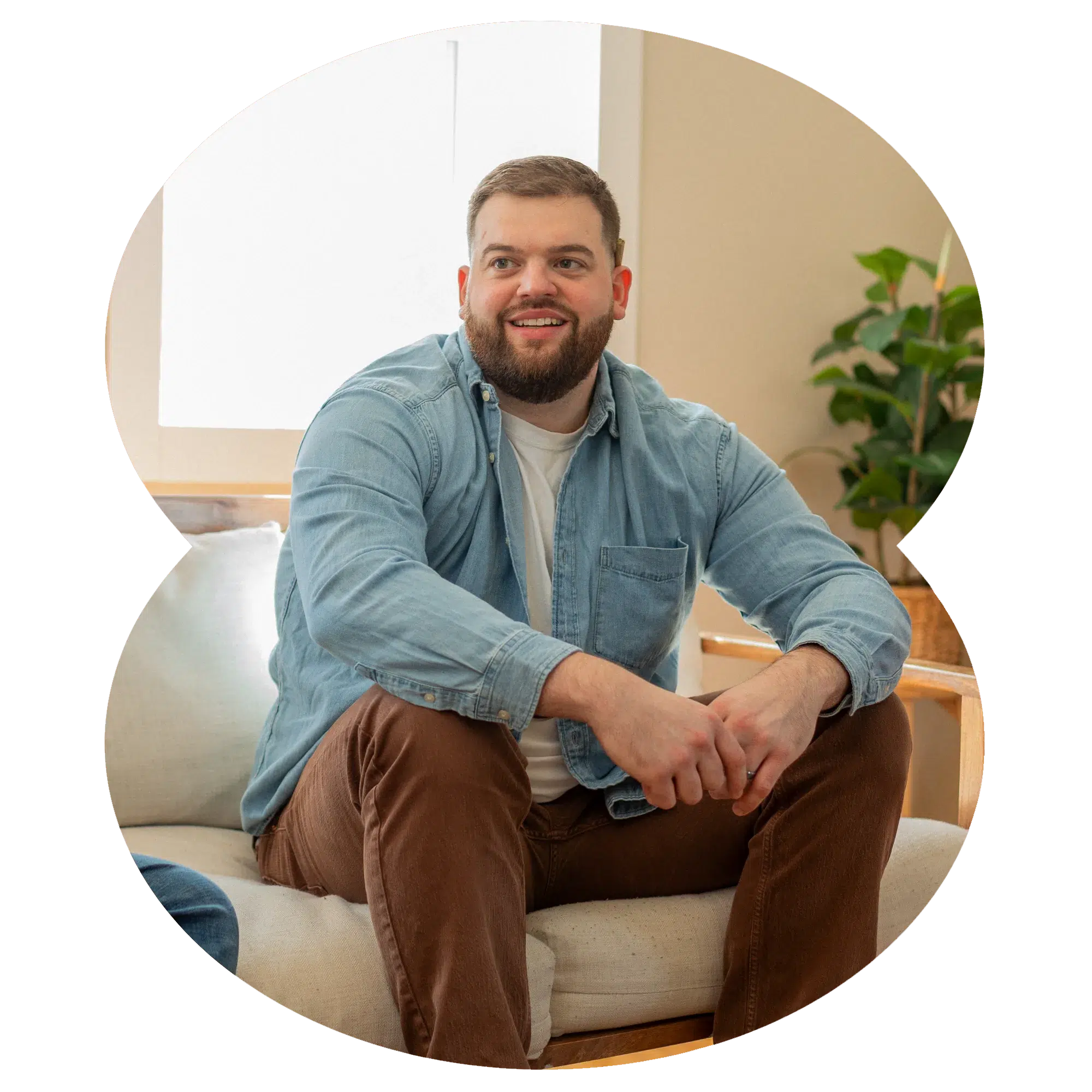 A bearded man in a denim shirt and brown pants sits on a local dispensary couch, looking away from the camera.